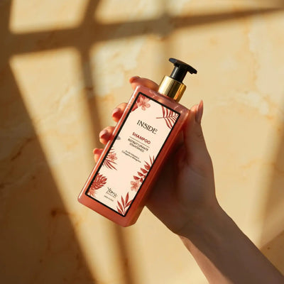 Una bottiglia rettangolare rosa di shampoo idratante per interni con dispenser a pompa nero e oro, con un'etichetta ornata da motivi di foglie rosse.