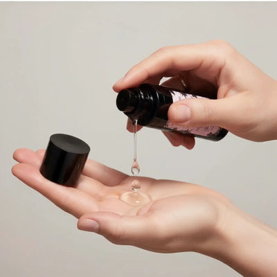 Una elegante bottiglia nera con finitura opaca e etichetta floreale rosa delicata, tenuta mentre gocce di liquido chiaro colano dal suo tappo nel palmo aperto.